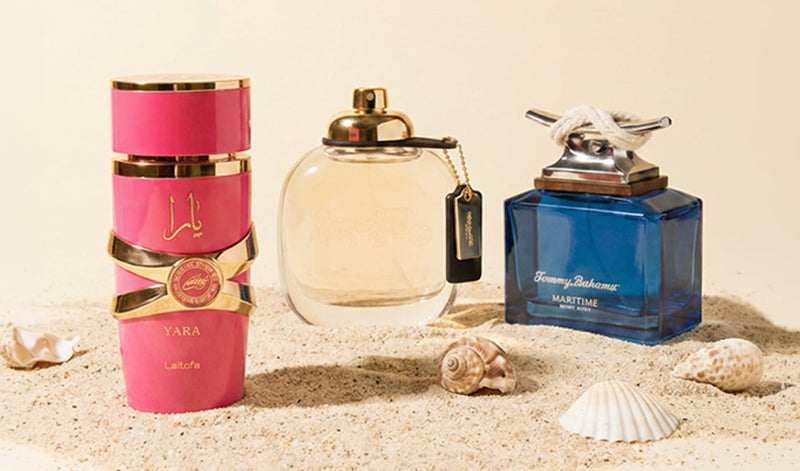 Three perfume bottles—pink Yara, clear Coach, and blue Tommy Bahama Maritime—displayed on sand with seashells.