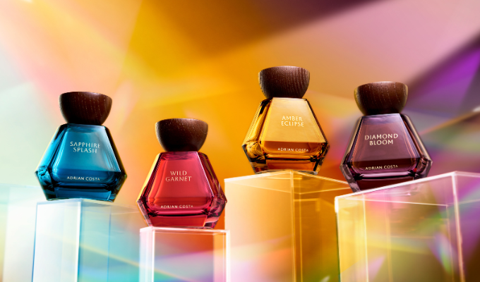 Four colorful Adrian Costa perfume bottles—Sapphire Splash, Wild Garnet, Amber Eclipse, and Diamond Bloom—displayed on clear pedestals against a soft, vibrant abstract background.