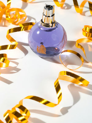 A round lavender-colored perfume bottle with a silver cap, surrounded by festive gold ribbon, representing a budget-friendly fragrance option.