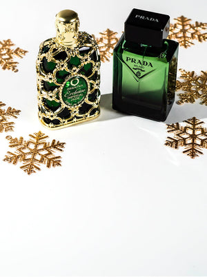Two green holiday-themed perfume bottles — Orientica’s Exclusive Imperial Jade in an ornate gold design and Prada Milano’s sleek green glass — surrounded by glittering gold snowflakes on a white backdrop.