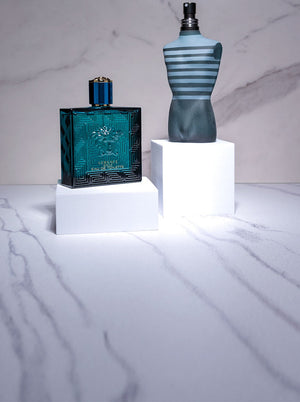Versace Eros and Jean Paul Gaultier Le Male fragrances displayed on white pedestal blocks against a marble background.