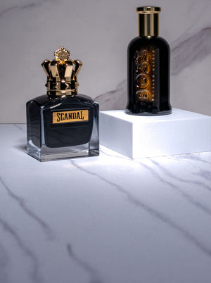 Jean Paul Gaultier Scandal and Boss Bottled Elixir fragrances displayed on white pedestal blocks against a marble background.