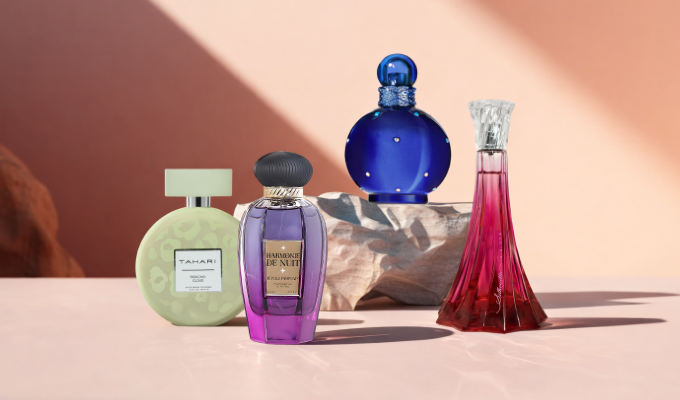 Four perfume bottles displayed on a smooth surface with a warm, desert-toned background, including a pale green Tahari bottle, a purple faceted bottle with a black cap, a round cobalt blue bottle resting on a stone, and a tall red gradient bottle with a clear decorative top.