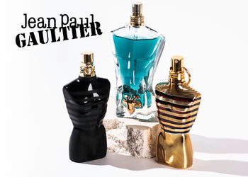 Jean Paul Gaultier fragrances displayed in signature torso-shaped bottles, including black, gold, and teal versions, set on a white background with stone props. 6