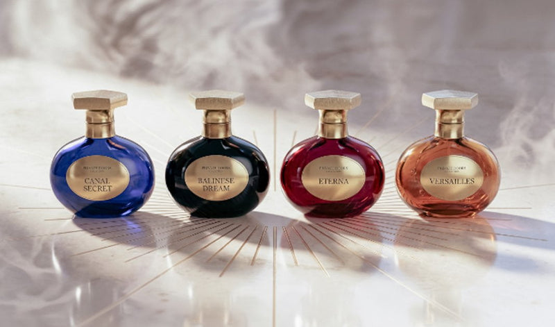 Four ornate perfume bottles in blue, black, red, and rose-gold displayed side by side on a luminous marble surface with a soft, elegant background.