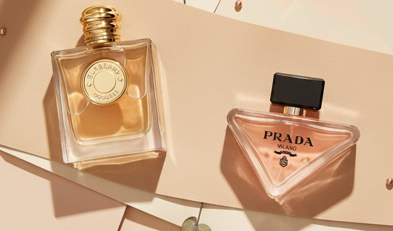 Two luxury perfume bottles, including Prada and  Burberry fragrances, arranged on a beige background with warm sunlight and soft shadows